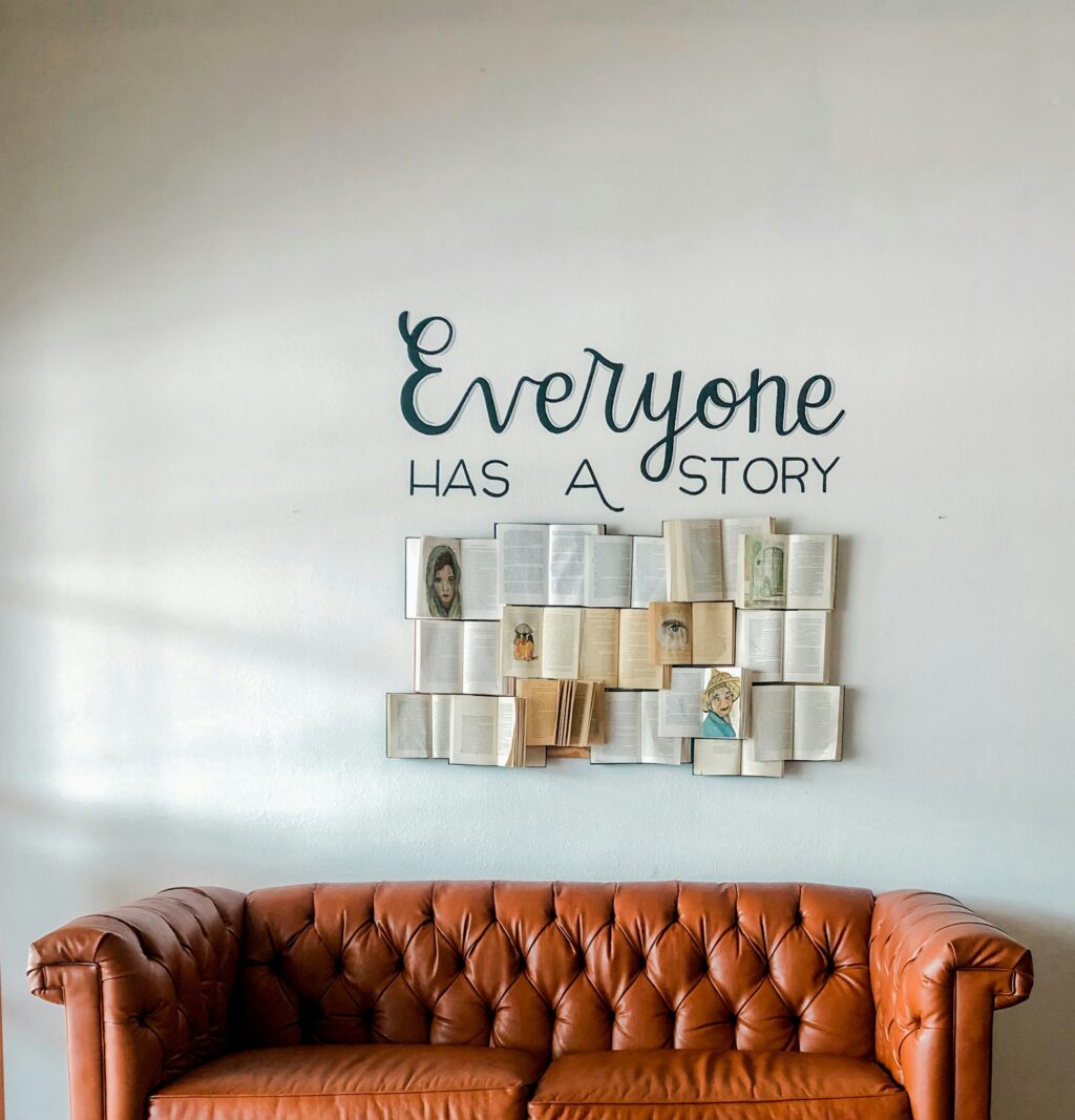 Schriftzug „Everyone has a story“ an einer Wand über aufgeschlagenen Büchern, davor ein braunes Ledersofa